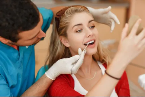Dentist examining a smiling young woman's teeth as she looks at her reflection in a handheld mirror