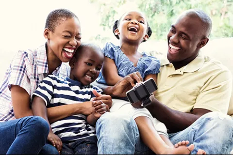 Happy family of four laughing and enjoying time together with a smartphone on a bright day.