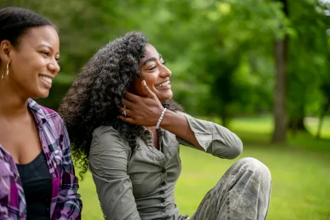 Two young women smiling and enjoying a sunny day sitting on grass in a green park outdoors.