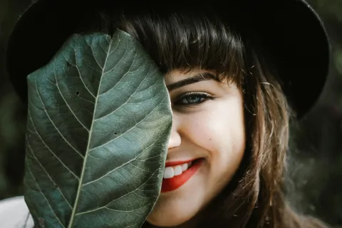Smiling woman with red lipstick and black hat holding a large green leaf covering half her face outdoors.