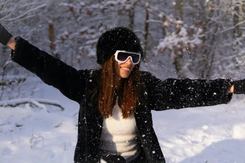 Joyful woman wearing winter clothes and sunglasses enjoying falling snow outdoors in a snowy forest.