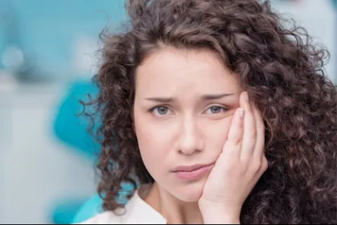 Young woman with curly hair holding her cheek, expressing pain or discomfort in a soft-focused indoor setting.