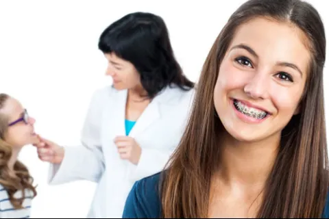 Smiling teenage girl with braces in foreground and orthodontist examining a young patient in the background.