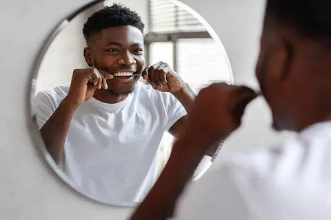 Smiling young man flossing teeth in front of round mirror wearing white t-shirt in bright room
