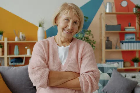 Smiling elderly woman in pink sweater posing with arms crossed in colorful cozy living room with shelves and cushions.