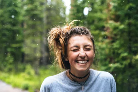 Young woman smiling with eyes closed outdoors in forest with rain droplets falling around her.