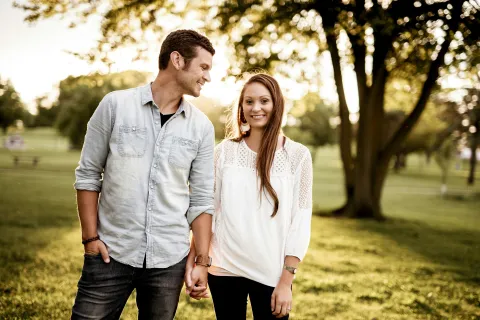 Couple holding hands walking in park at sunset with smiles and greenery in background