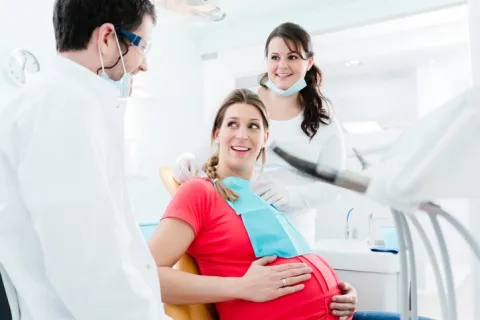 Pregnant woman smiling in dental chair with dentist and hygienist in bright dental office setting.