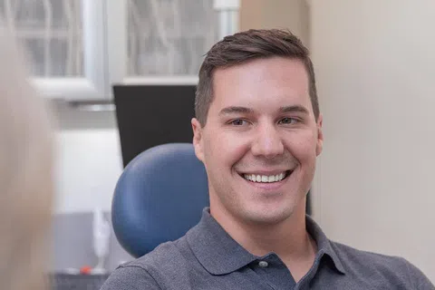 Smiling man sitting in a dental chair wearing a gray polo shirt in a bright dental office
