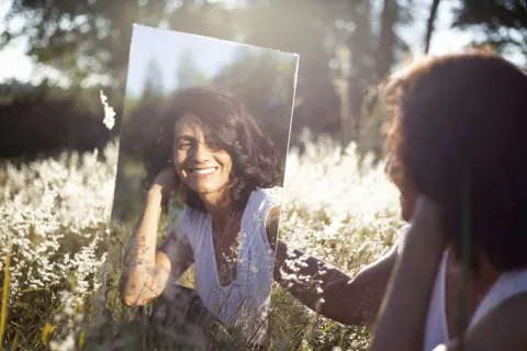 Woman smiling at her reflection in a mirror while sitting in a sunlit field of tall grass and wildflowers.