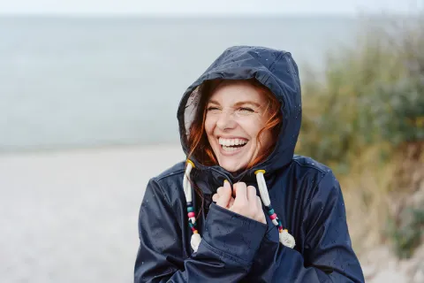 A woman in a rain jacket smiles joyfully by the beach on a rainy day.