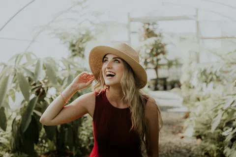 Smiling woman in a straw hat and red dress enjoys a sunny day inside a lush greenhouse filled with greenery.