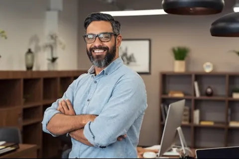 Smiling bearded man with glasses and light blue shirt stands confidently in modern office space.