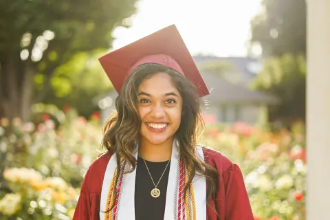 Smiling young woman in maroon graduation gown and cap with salutatorian stole outdoors in sunlight