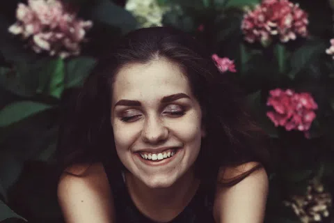 Young woman with closed eyes smiling surrounded by green leaves and pink flowers in soft natural light