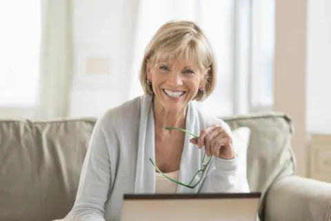 Smiling older woman using a laptop at home, sitting on a sofa with glasses in hand.