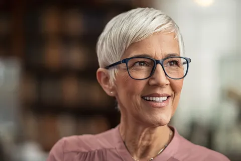 Smiling older woman with short silver hair and glasses in a warm, inviting setting.