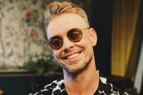 Smiling young man wearing round sunglasses and a black and white patterned shirt indoors with decorative background.