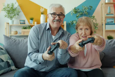 Senior couple smiling and playing video games together on a couch in a colorful living room.