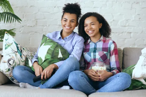 Two smiling young women sitting on a couch with tropical-themed pillows, enjoying popcorn and time together