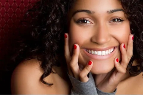 Close-up of a smiling woman with curly hair and red nail polish holding her face against a patterned background