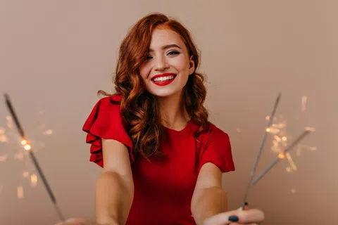Smiling red-haired woman in red dress holding sparklers against neutral background