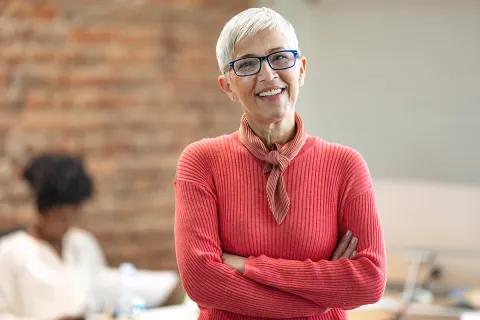 Smiling older woman in a sweater, arms crossed, standing in an office setting with a blurred background.