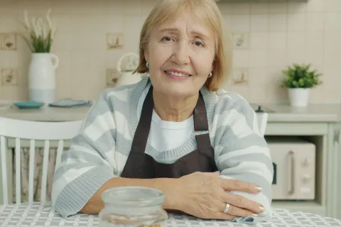 Smiling elderly woman wearing apron sitting at kitchen table with jar and plants in background