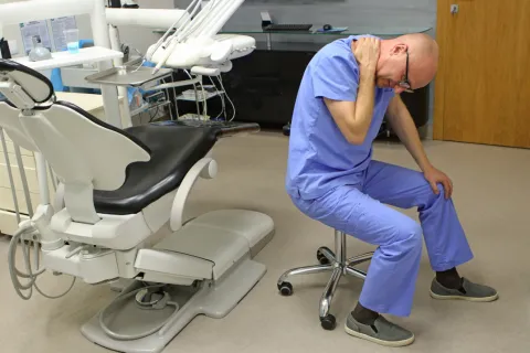 Dentist in blue scrubs sitting on a stool holding his neck in pain next to a dental chair in clinic.