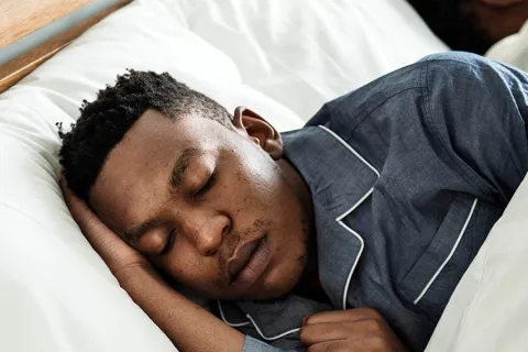A young man peacefully sleeping in bed, wearing blue pajamas, with another person asleep nearby.