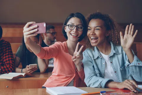 Two female students smiling and taking a selfie in a classroom with other students studying in the background