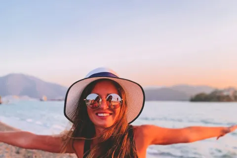 Smiling woman in hat and sunglasses enjoying sunset on rocky beach with arms outstretched by the sea.