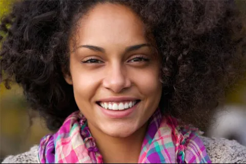Smiling woman with curly hair wearing a colorful scarf in a natural outdoor setting.