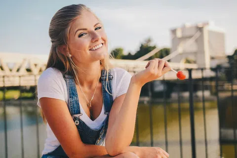 Smiling young woman in denim overalls holding a red lollipop outdoors by a railing near water at sunset.