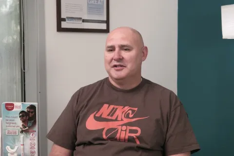 Man in brown Nike Air t-shirt sitting in a room with dental product boxes and a framed certificate on the wall.