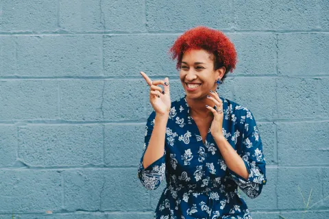 Smiling woman with red curly hair wearing a blue patterned dress pointing to the side against a gray brick wall.