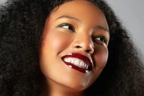 Smiling woman with natural curly hair wearing glossy dark red lipstick and gold eyeshadow on a gray background