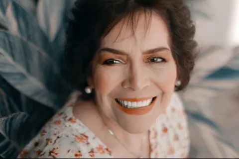 Smiling older woman with short hair, wearing a floral dress against a leafy backdrop.