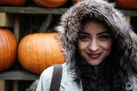 Woman in a fur-lined hooded coat smiles with pumpkins displayed on wooden shelves behind her.