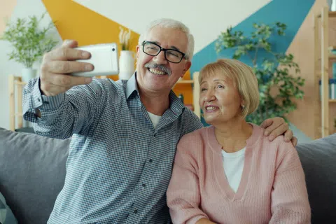 Smiling elderly couple sitting on couch taking a selfie with smartphone in a colorful living room