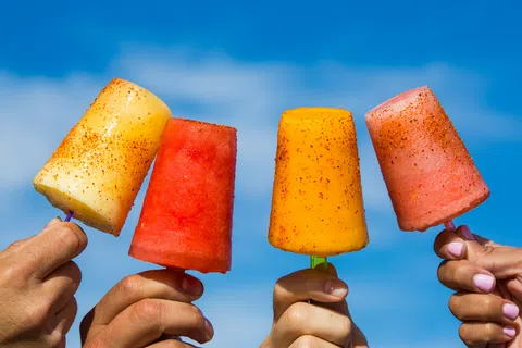 Four hands holding colorful fruit popsicles sprinkled with chili powder against a bright blue sky.