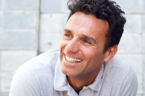 Smiling man with curly hair in a gray polo shirt against a textured wall background.