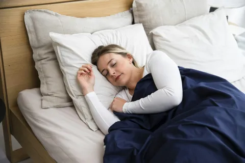 Woman peacefully sleeping on white pillows in a wooden bed with beige sheets and a navy blue blanket.