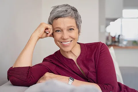 Smiling mature woman with short gray hair relaxing on a couch wearing a burgundy top and silver watch.
