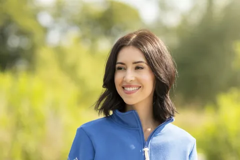 Smiling woman in blue and navy color-block pullover sweatshirt standing outdoors by water and greenery.