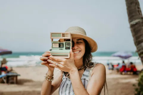 Young woman in a hat smiling and holding a vintage instant camera on a sunny beach with ocean in background