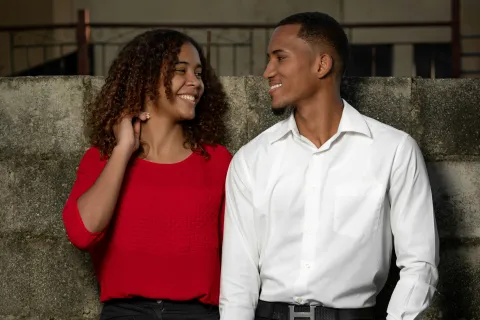 Smiling young couple holding hands, leaning against a concrete wall, dressed in red top and white shirt.