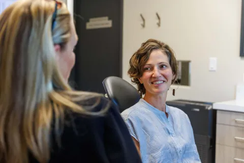 Two women in conversation in a modern office setting, one smiling and seated in a chair facing the other.