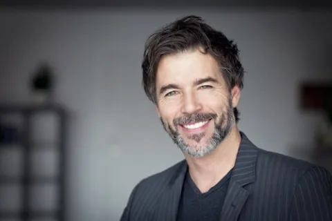 Smiling mature man with gray beard wearing a dark blazer and black shirt in a professional setting.