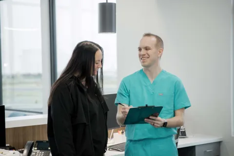 Healthcare professional in teal scrubs discussing information with woman in black in a bright modern office.